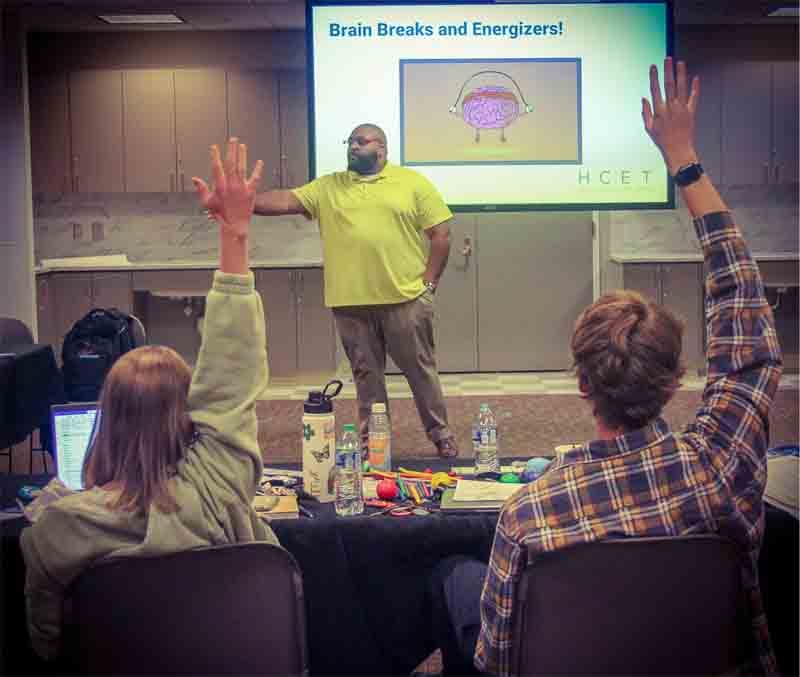 A dark-skinned man with a beard, wearing a green shirt. He is in a kitchen with a projector. In front of him, there are two young people raising their hands to ask something about Brain Breaks and Energizers.