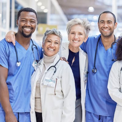 5 people in a hospital, 2 of them are nurses wearing blue nurse uniforms, and 3 of them are female doctors, each in a white coat. They are all hugging while smiling. One of them is holding a list in her hand.
